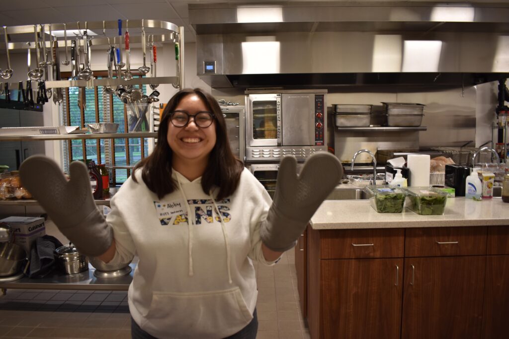 a young woman waves at the camera with both hands covered in oven mits as she stands in a church kitchen