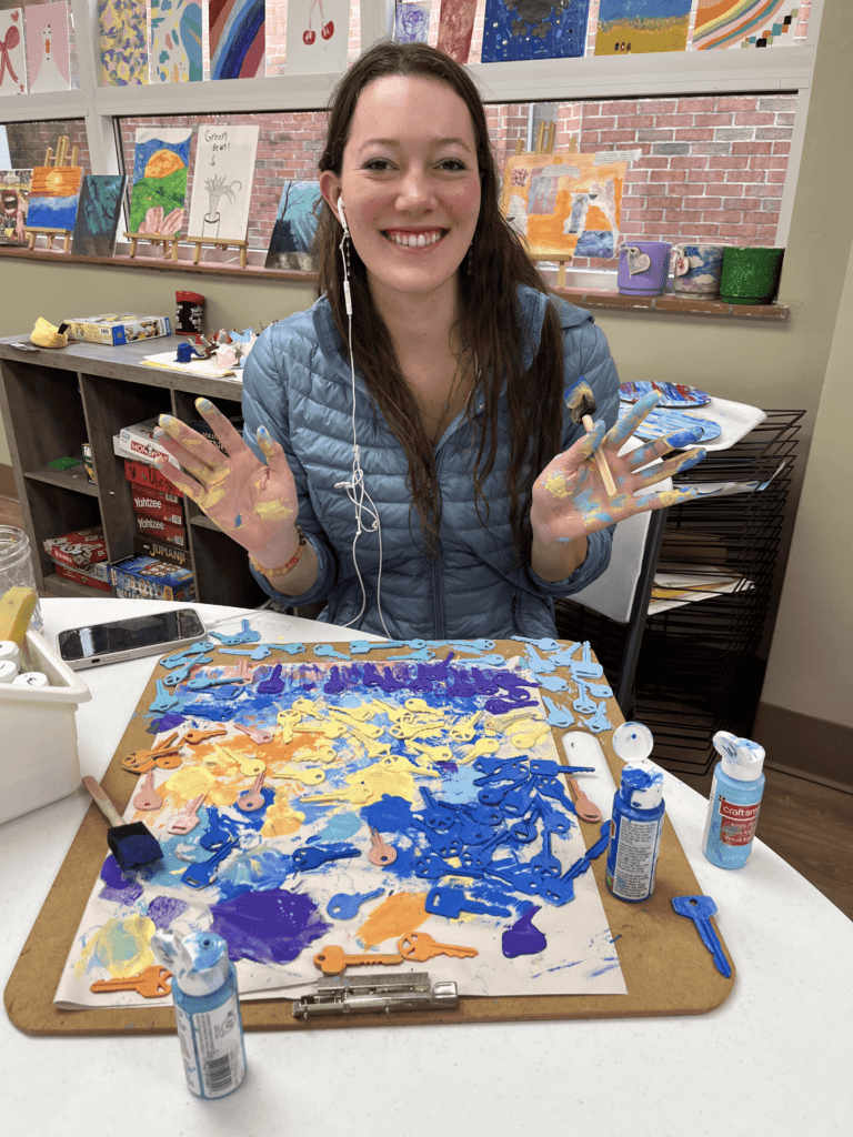 A smiling intern shows off her hands, which are covered in paint. In front of her sits a colorful assortment of hand-painted keys. 